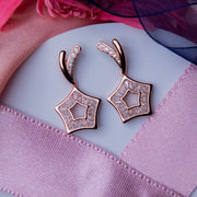 A pair of rose gold-colored earrings with a star-shaped design, featuring multiple points and adorned with cubic zirconia, displayed on a pink and white fabric surface.